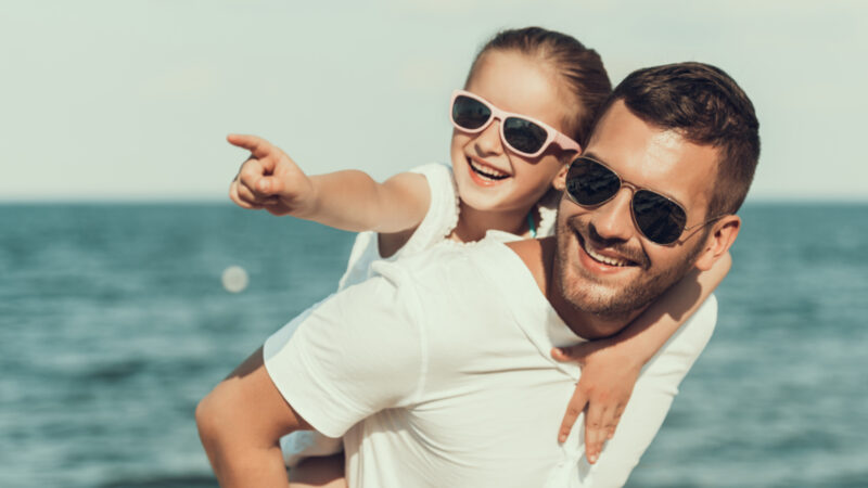 ein kleines Mädchen wird auf dem Rücken ihres Vaters getragen, beide tragen Sonnenbrillen und lächeln in die Kamera